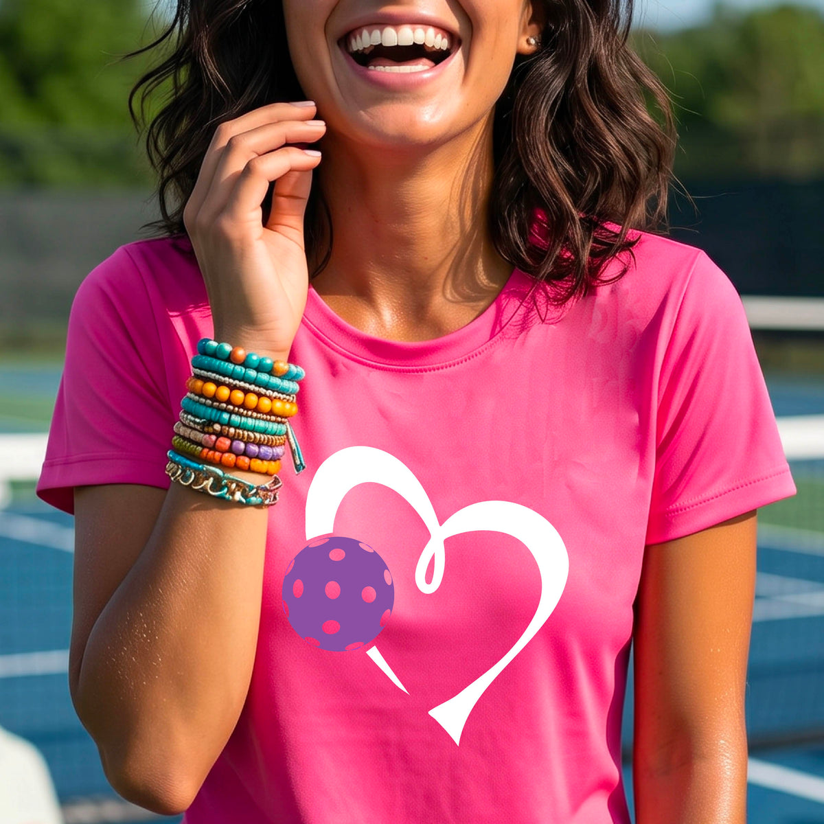 A woman wearing a bright pink short-sleeve t-shirt with a white heart outline and a purple pickleball graphic on the front, set against a blurred pickleball court background.
