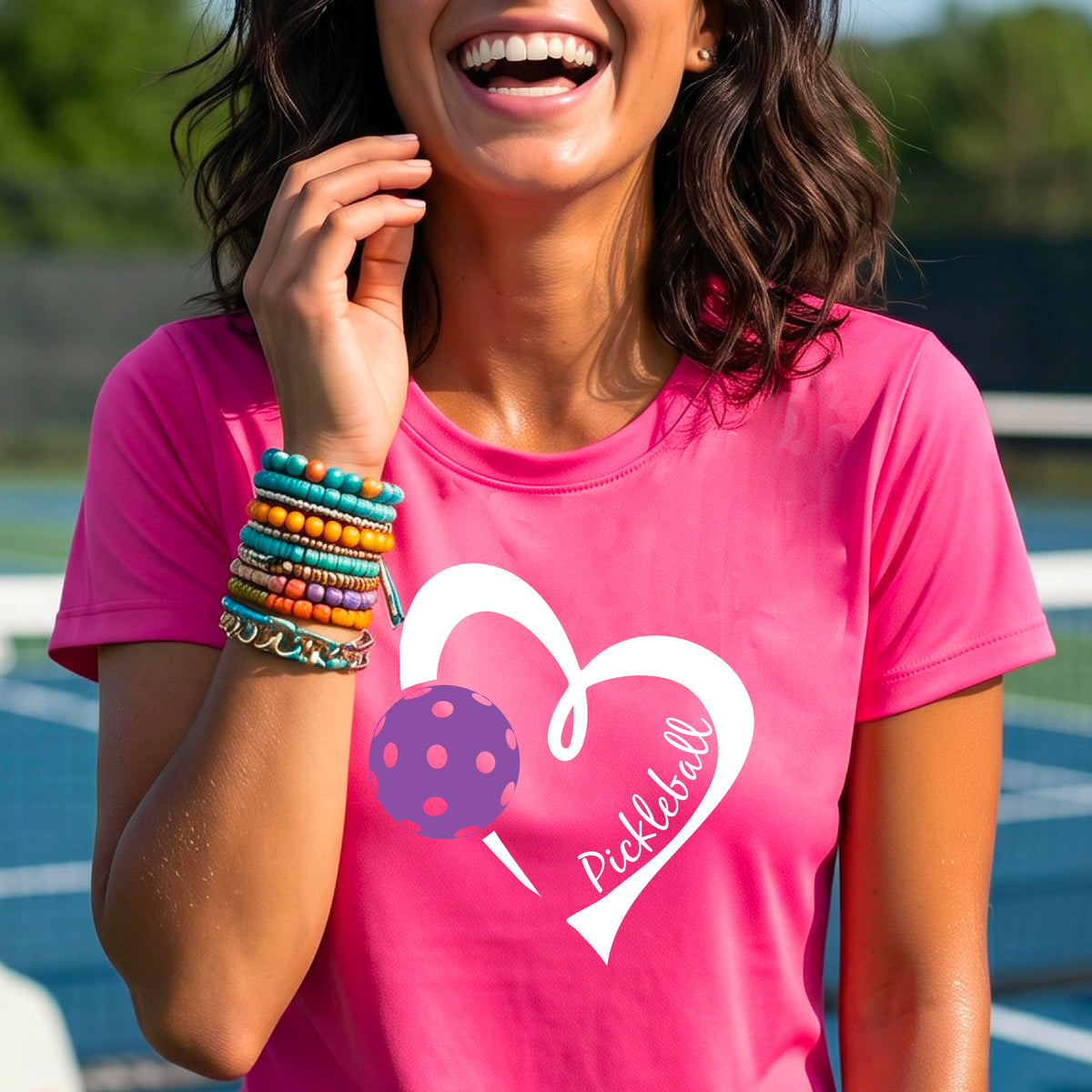 A woman wearing a pink graphic t-shirt with a white heart outline, a purple pickleball, and the word 'Pickleball' printed on the front, set against a blurred tennis court background.