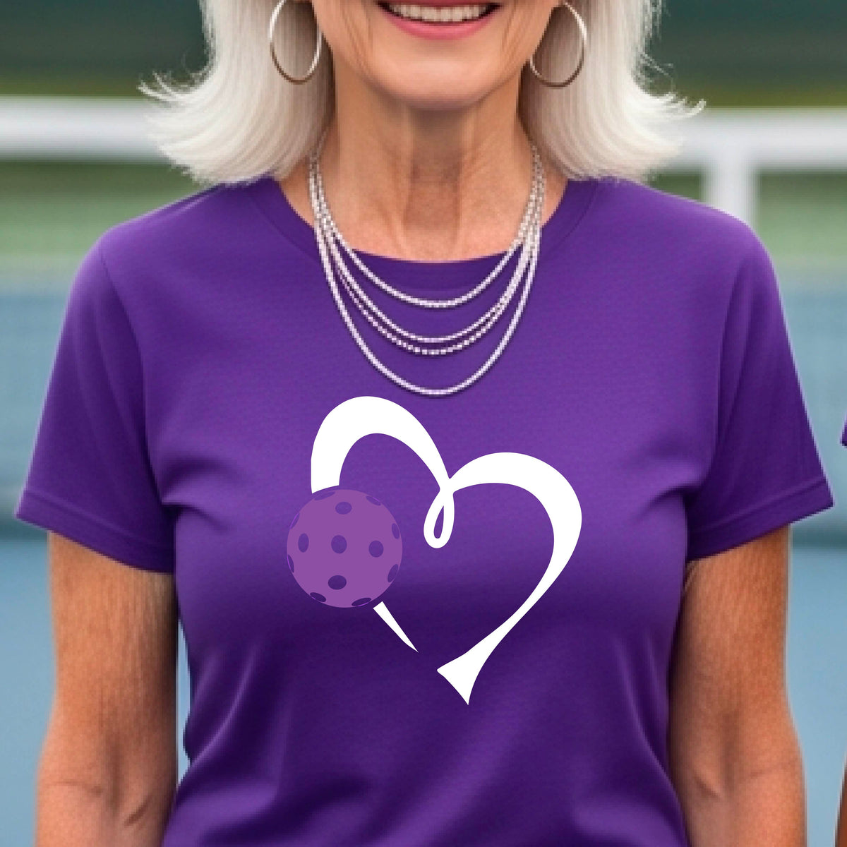 A woman wearing a purple short-sleeve t-shirt with a white heart outline and a purple pickleball graphic on the front, set against a blurred pickleball court background.