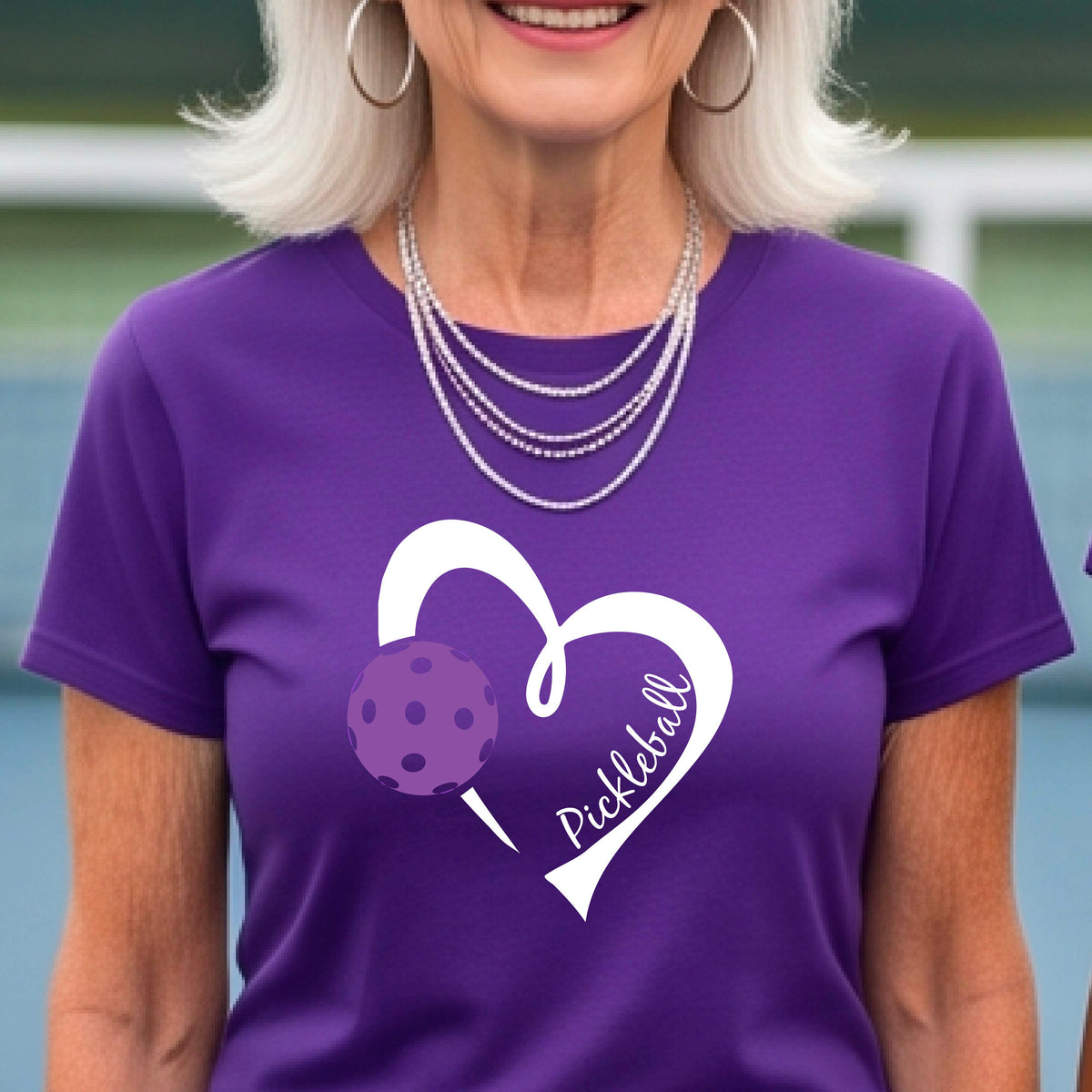 A woman wearing a purple graphic t-shirt with a white heart outline, a purple pickleball, and the word 'Pickleball' printed on the front, set against a blurred tennis court background.
