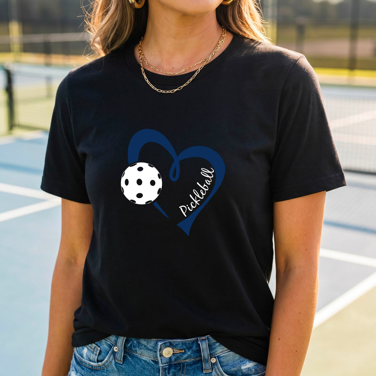 A woman wearing a black graphic t-shirt with a navy blue heart, a white pickleball, and the word 'Pickleball' written on the front,.