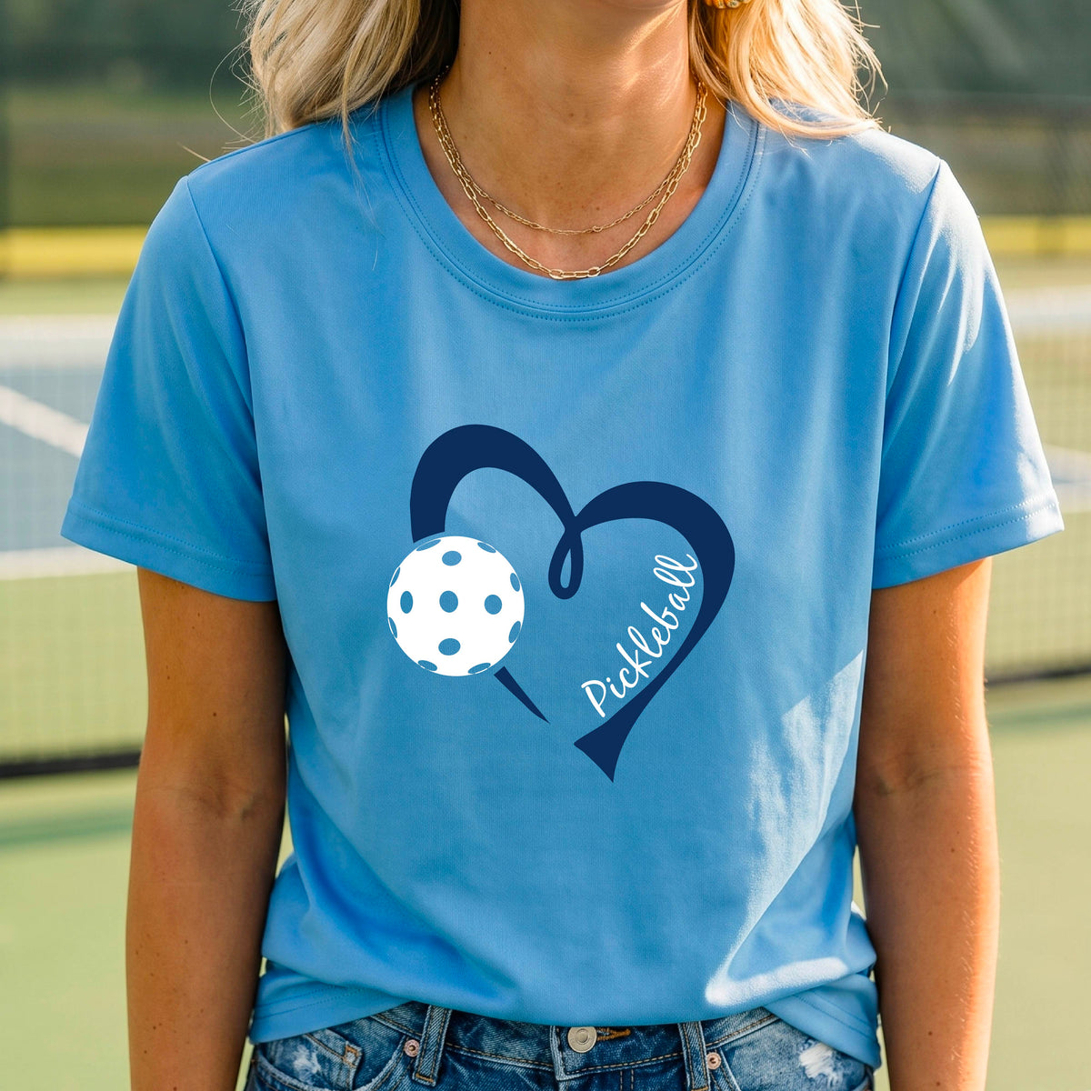 A woman wearing a bright blue  graphic t-shirt with a navy blue heart, a white pickleball, and the word 'Pickleball' written on the front,.