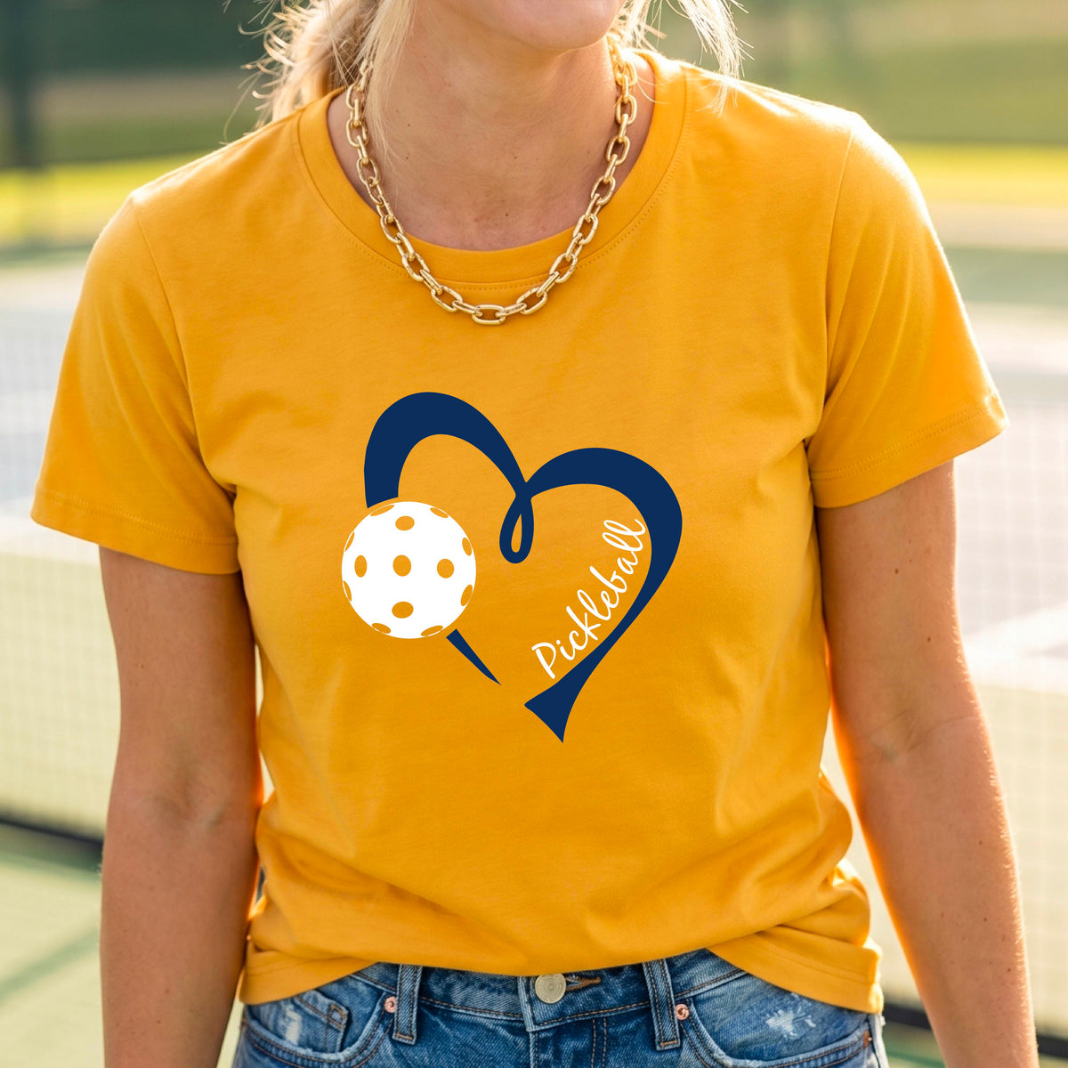 A woman wearing a bright yellow graphic t-shirt with a navy blue heart, a white pickleball, and the word 'Pickleball' written on the front,.