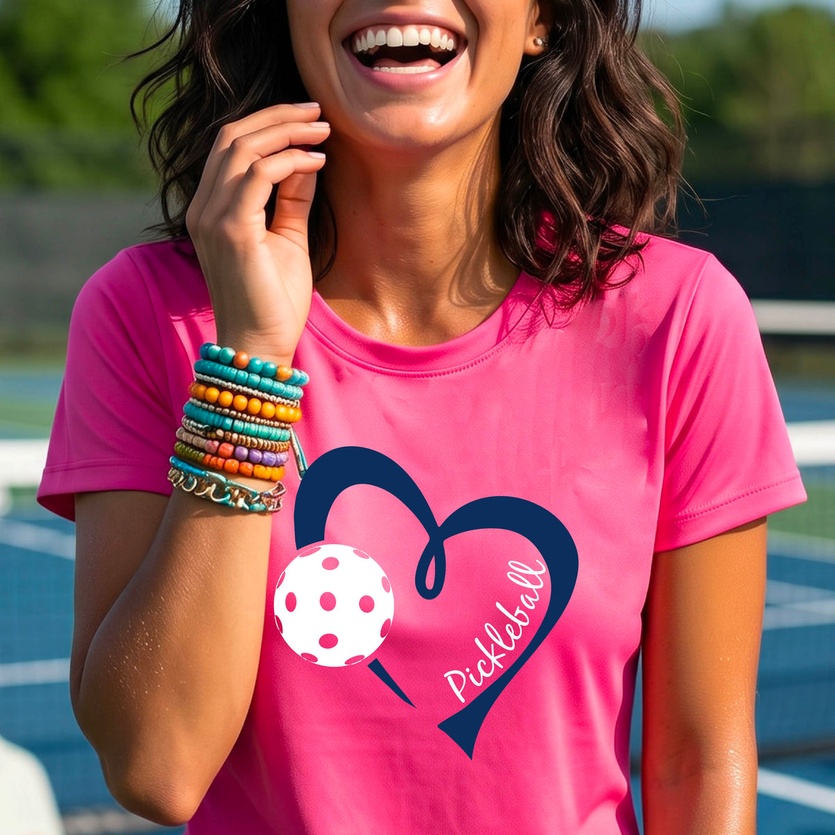 A woman wearing a bright pink graphic t-shirt with a navy blue heart, a white pickleball, and the word 'Pickleball' written on the front,.