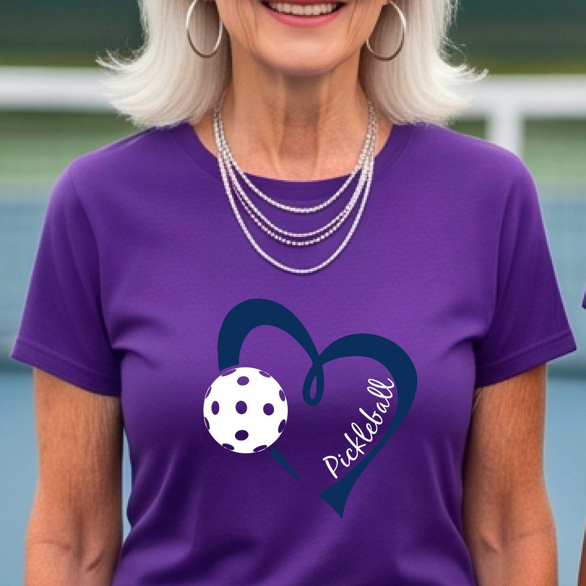 A woman wearing a purple graphic t-shirt with a navy blue heart, a white pickleball, and the word 'Pickleball' written on the front,.