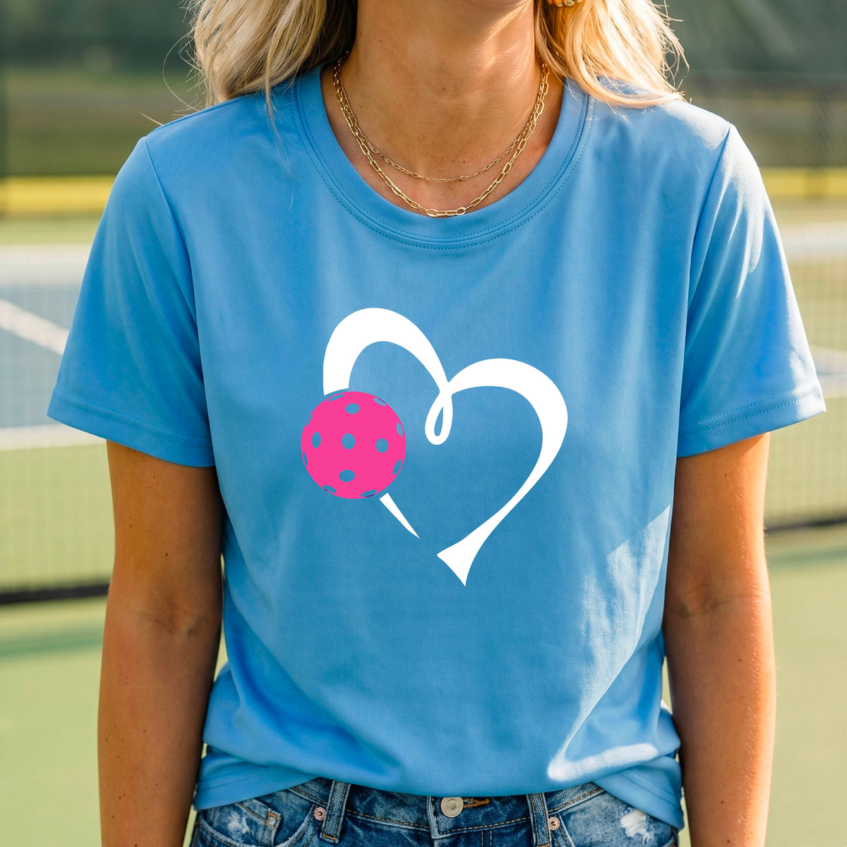 A woman wearing a light blue t-shirt with a white heart outline and a pink pickleball graphic on the front, set against a blurred pickleball court background.