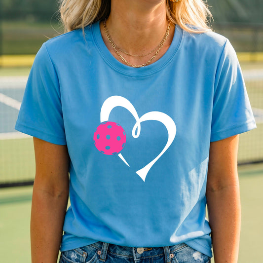 A woman wearing a light blue t-shirt with a white heart outline and a pink pickleball graphic on the front, set against a blurred pickleball court background.