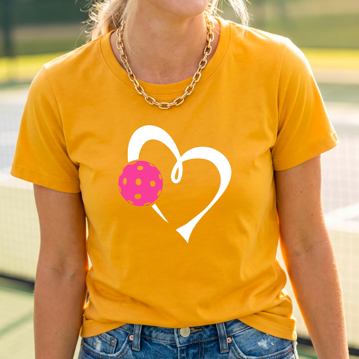 A woman wearing a mustard yellow gold t-shirt with a white heart outline and a pink pickleball graphic on the front, set against a blurred pickleball court background.