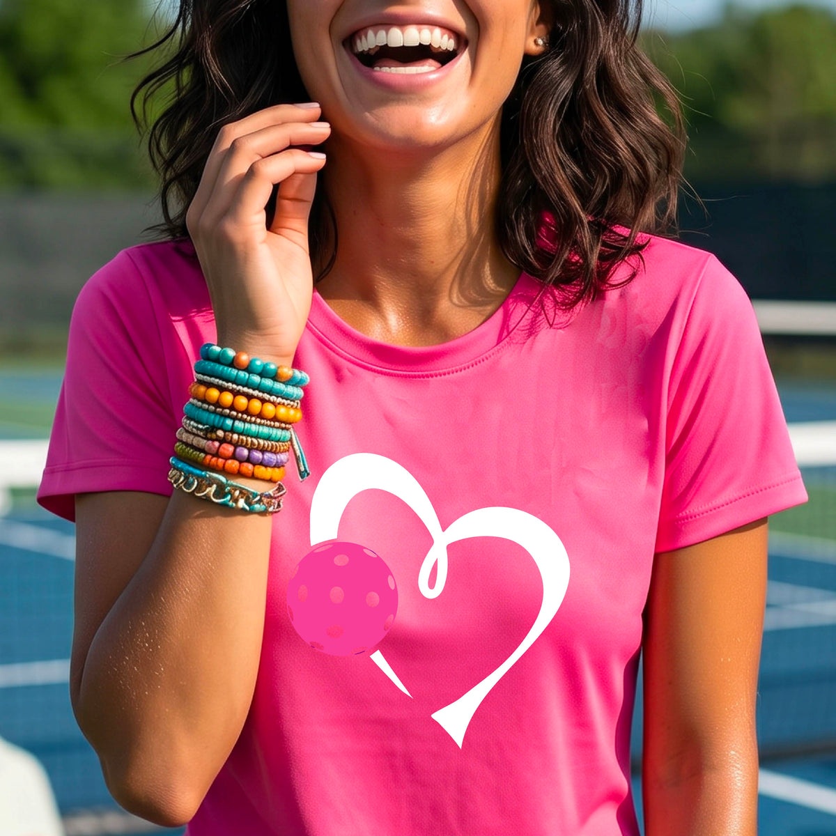 A woman wearing a pink t-shirt with a white heart outline and a pink pickleball graphic on the front, set against a blurred pickleball court background.