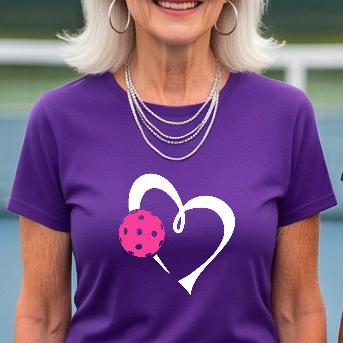 A woman wearing a purple t-shirt with a white heart outline and a pink pickleball graphic on the front, set against a blurred pickleball court background.