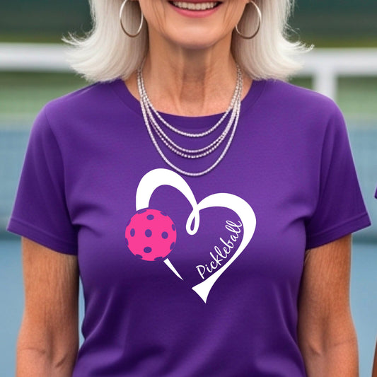 A woman wearing a purple t-shirt with a white heart outline, a pink pickleball graphic, and the word 'Pickleball' written inside the heart. The background is a blurred outdoor pickleball court.