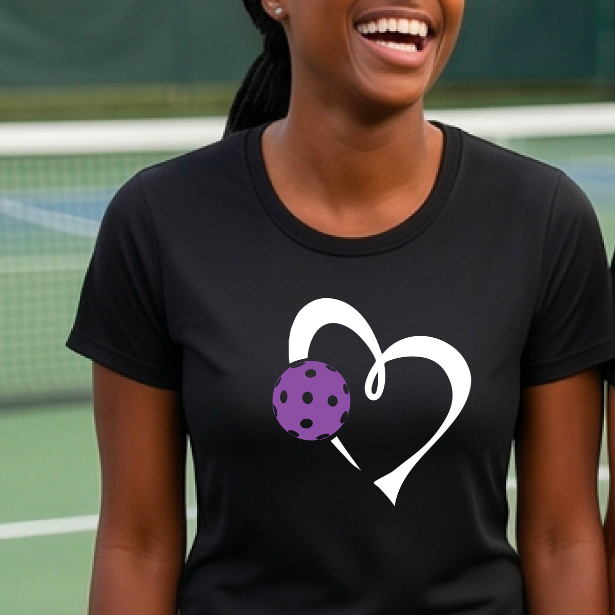 A woman wearing a black short-sleeve t-shirt with a white heart outline and a purple pickleball graphic on the front, set against a blurred pickleball court background.