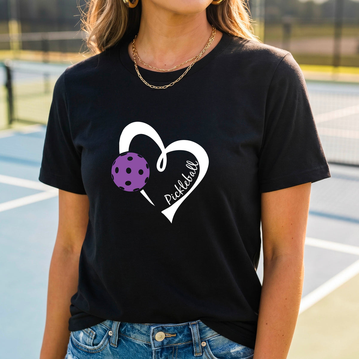 A woman wearing a black graphic t-shirt with a white heart outline, a purple pickleball, and the word 'Pickleball' printed on the front, set against a blurred tennis court background.