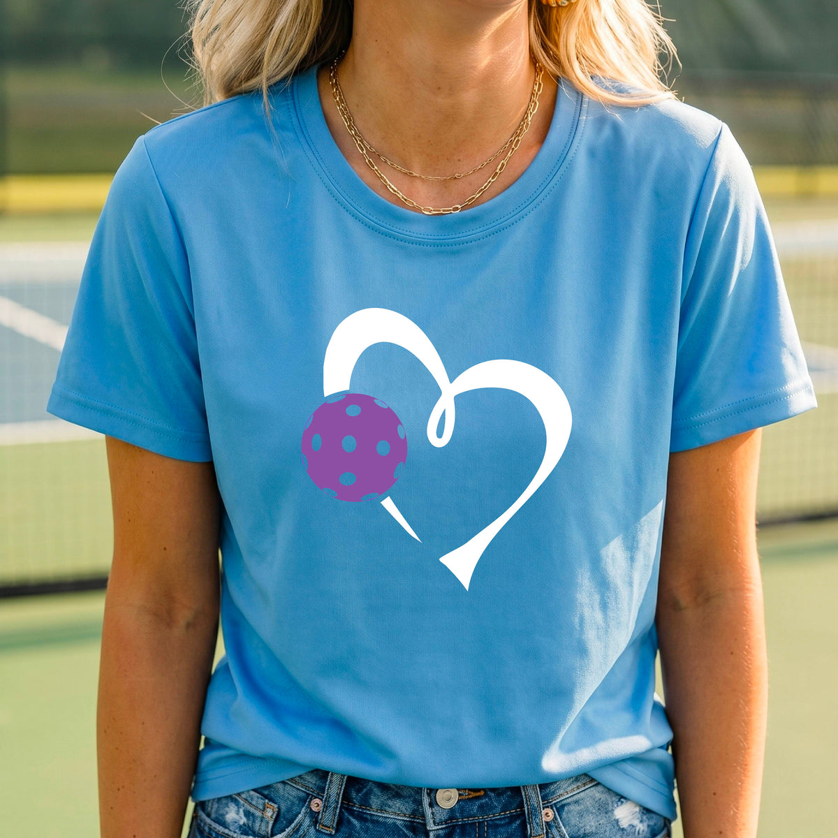 A woman wearing a blue short-sleeve t-shirt with a white heart outline and a purple pickleball graphic on the front, set against a blurred pickleball court background.