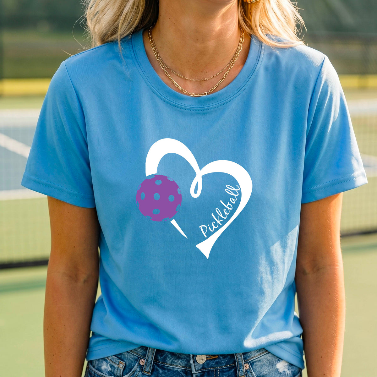 A woman wearing a bright blue graphic t-shirt with a white heart outline, a purple pickleball, and the word 'Pickleball' printed on the front, set against a blurred tennis court background.