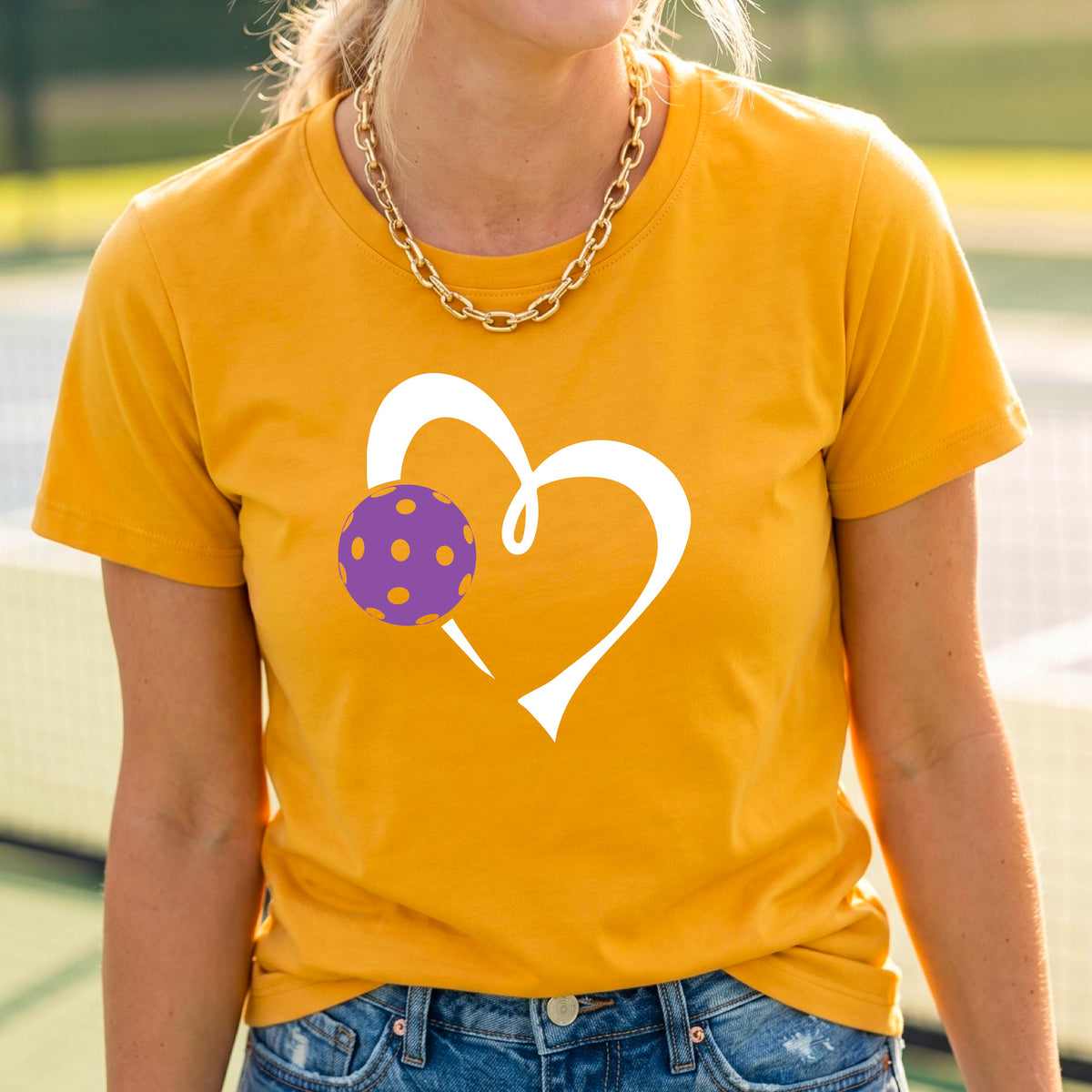A woman wearing a mustard yellow gold short-sleeve t-shirt with a white heart outline and a purple pickleball graphic on the front, set against a blurred pickleball court background.
