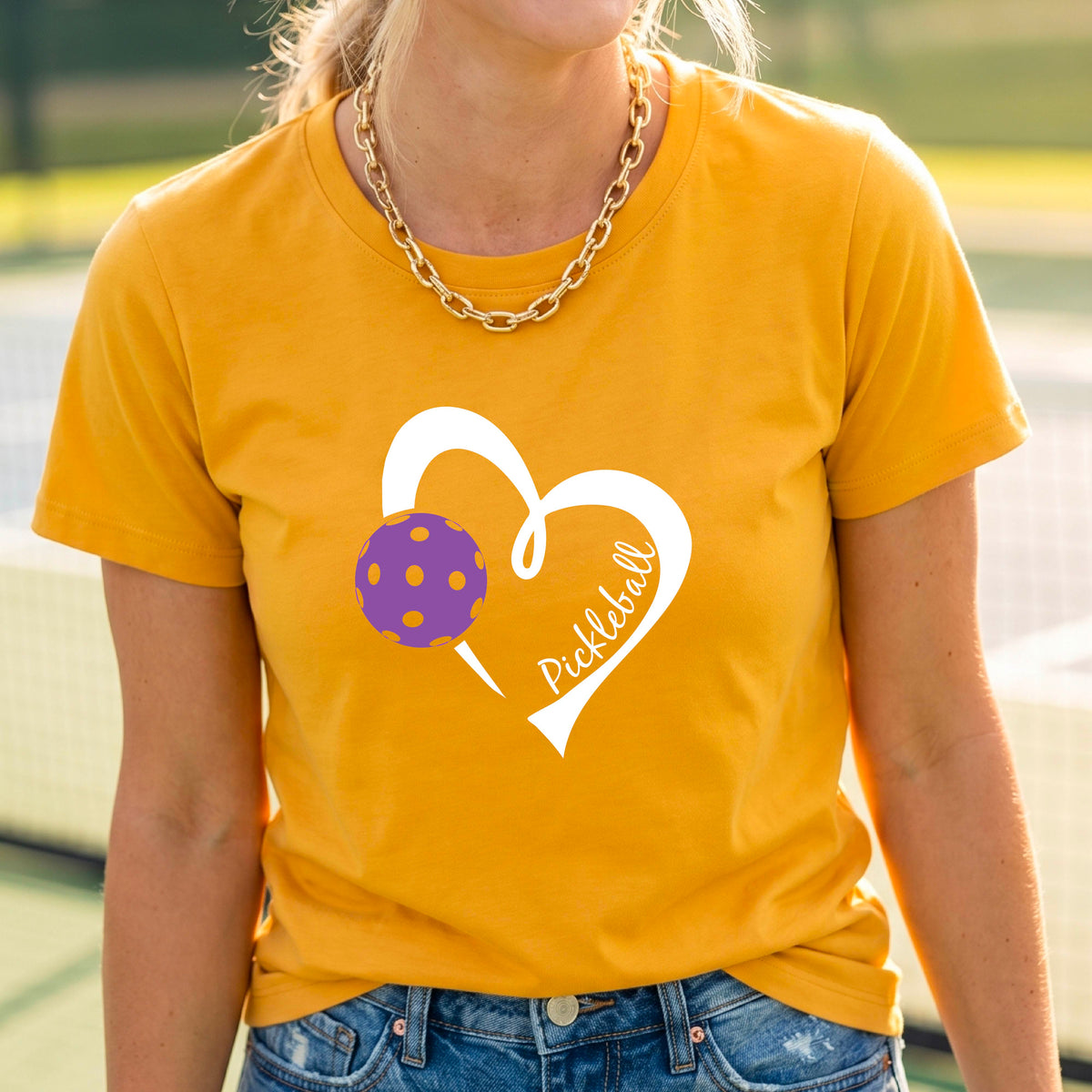 A woman wearing a mustard yellow gold graphic t-shirt with a white heart outline, a purple pickleball, and the word 'Pickleball' printed on the front, set against a blurred tennis court background.