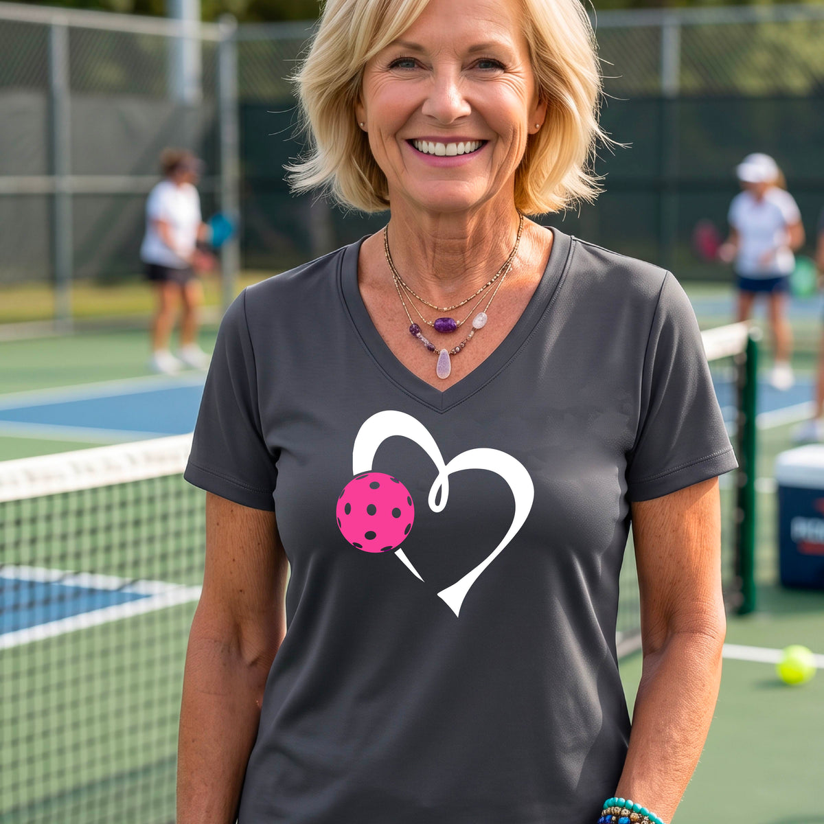 A woman laughing on a pickelball court wearing a dark gray V-neck t-shirt with a white heart outline and a pink pickleball graphic.
