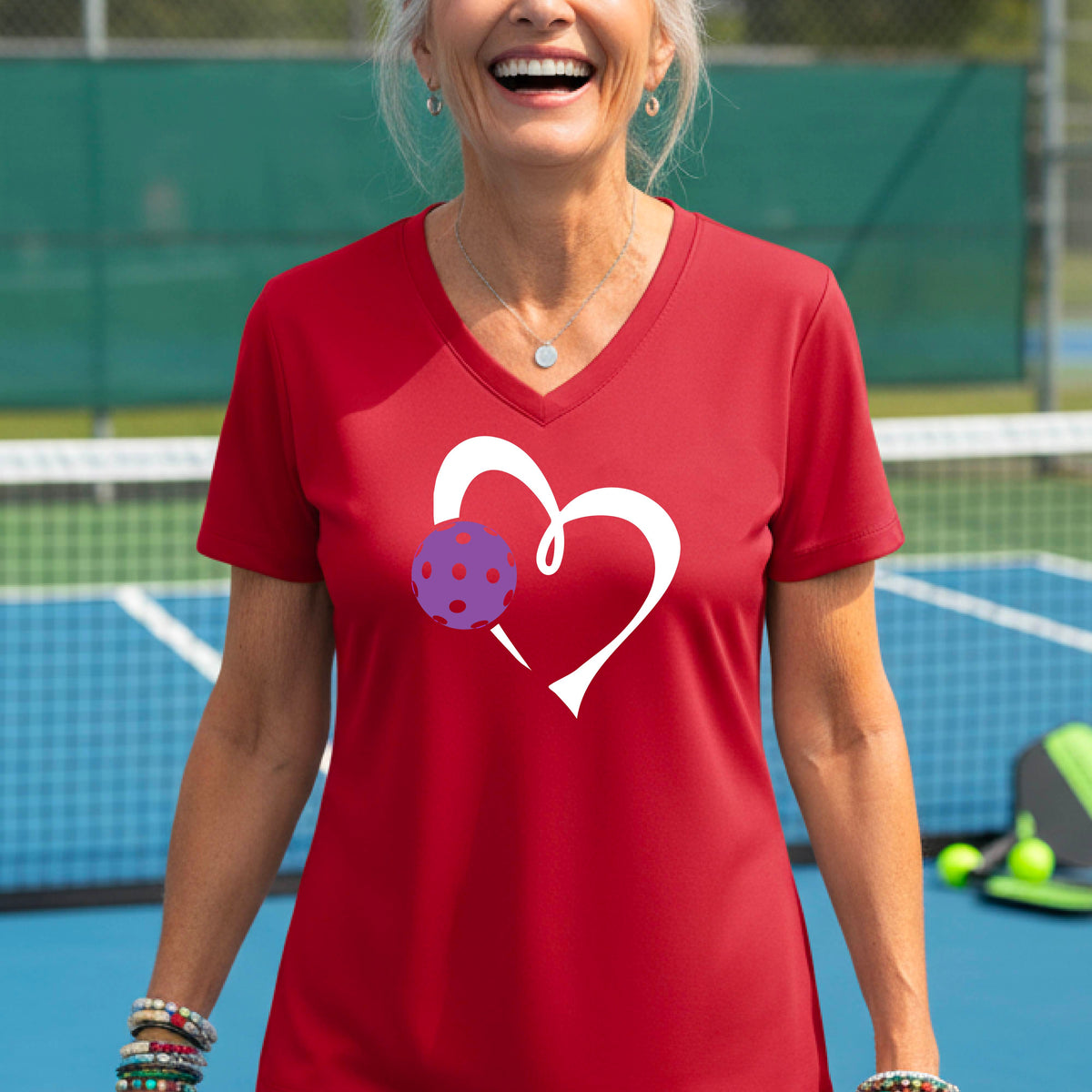 A woman wearing a red V-neck t-shirt with a white heart outline and a purple pickleball graphic, standing on a pickleball court.