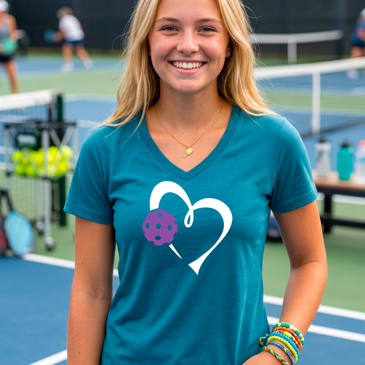 A woman wearing a teal V-neck t-shirt with a white heart outline and a purple pickleball graphic, standing on a pickleball court.