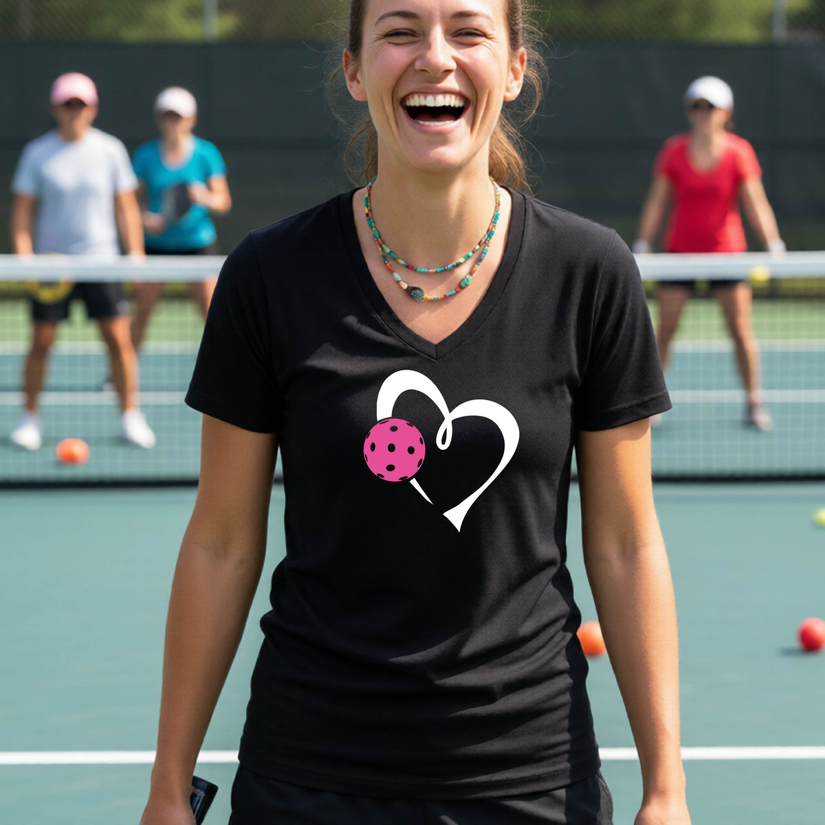 A woman laughing on a pickelball court wearing a black V-neck t-shirt with a white heart outline and a pink pickleball graphic.