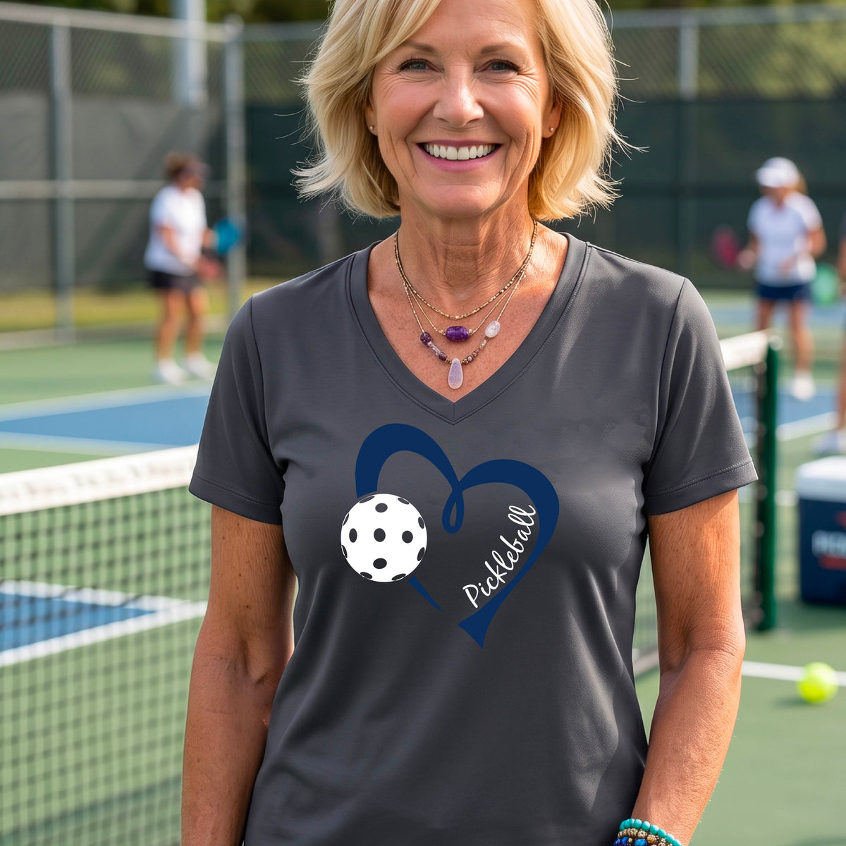 A woman on a pickleball court wearing a dark gray V-neck athletic t-shirt with a navy blue heart and pickleball graphic on the front.