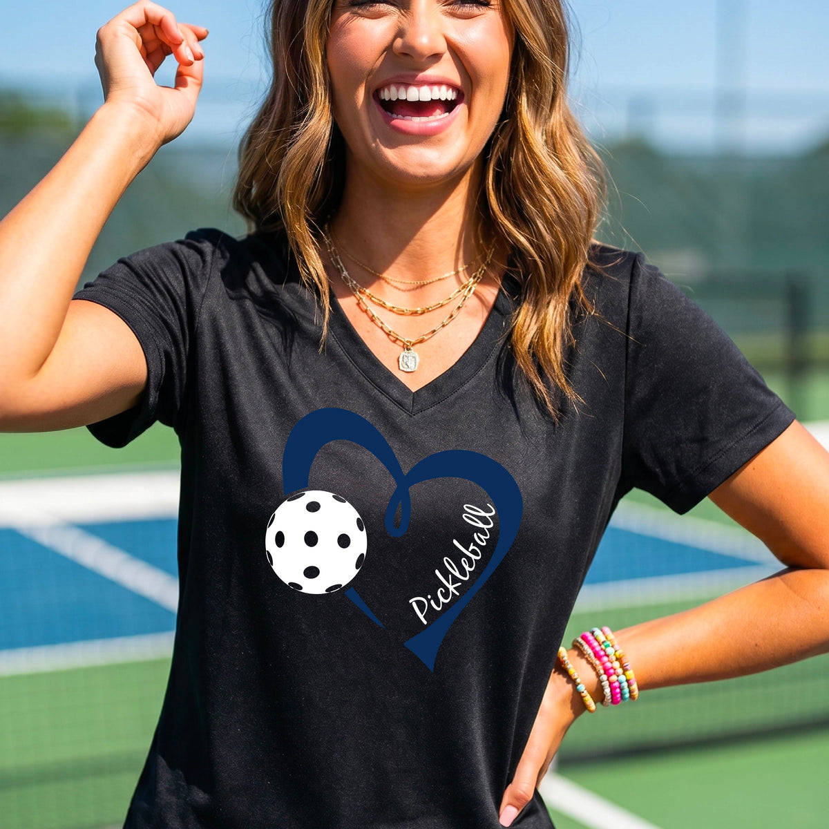 A woman on a pickleball court wearing a black V-neck athletic t-shirt with a navy blue heart and pickleball graphic on the front.