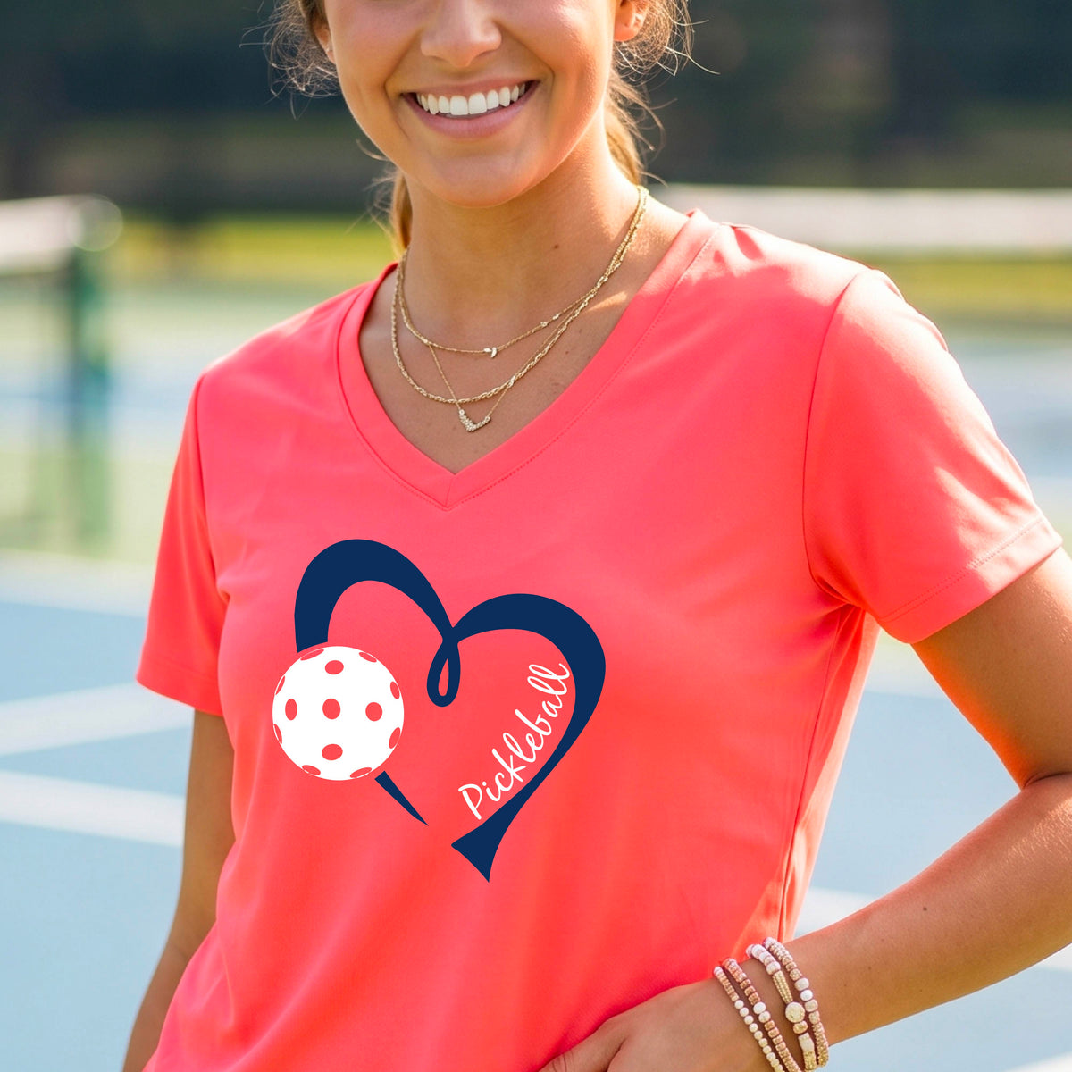 A woman on a pickleball court wearing a coral pink V-neck athletic t-shirt with a navy blue heart and pickleball graphic on the front.