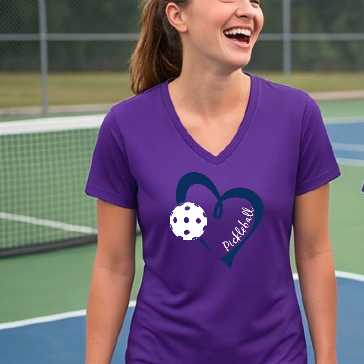 A woman on a pickleball court wearing a purple V-neck athletic t-shirt with a navy blue heart and pickleball graphic on the front.