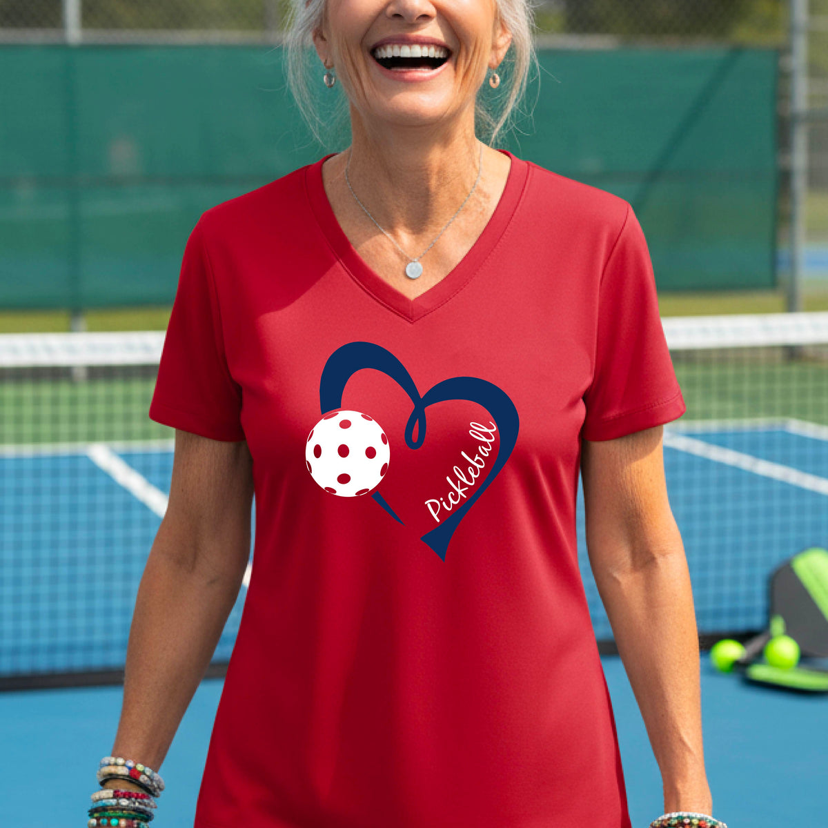 A woman on a pickleball court wearing a red V-neck athletic t-shirt with a navy blue heart and pickleball graphic on the front.