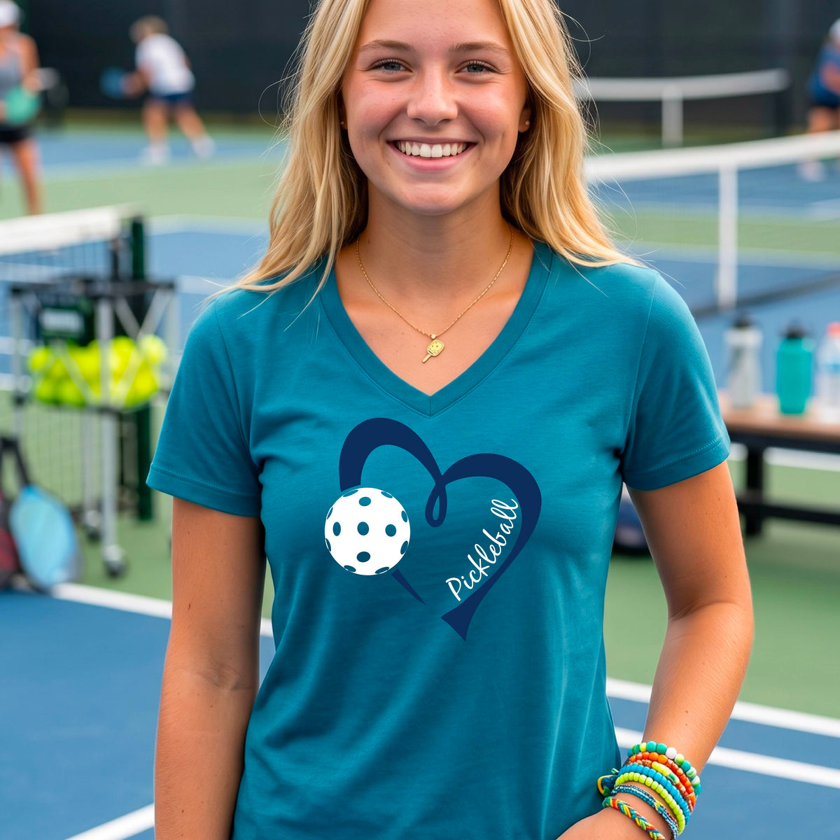 A woman on a pickleball court wearing a teal V-neck athletic t-shirt with a navy blue heart and pickleball graphic on the front.