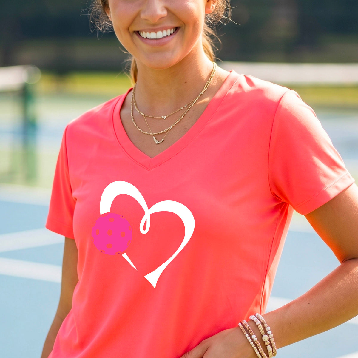 A woman laughing on a pickelball court wearing a coral pink V-neck t-shirt with a white heart outline and a pink pickleball graphic.