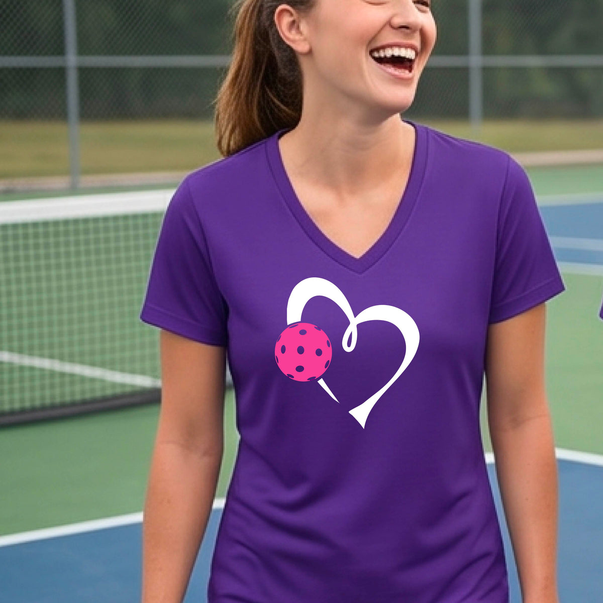 A woman laughing on a pickelball court wearing a purple V-neck t-shirt with a white heart outline and a pink pickleball graphic.
