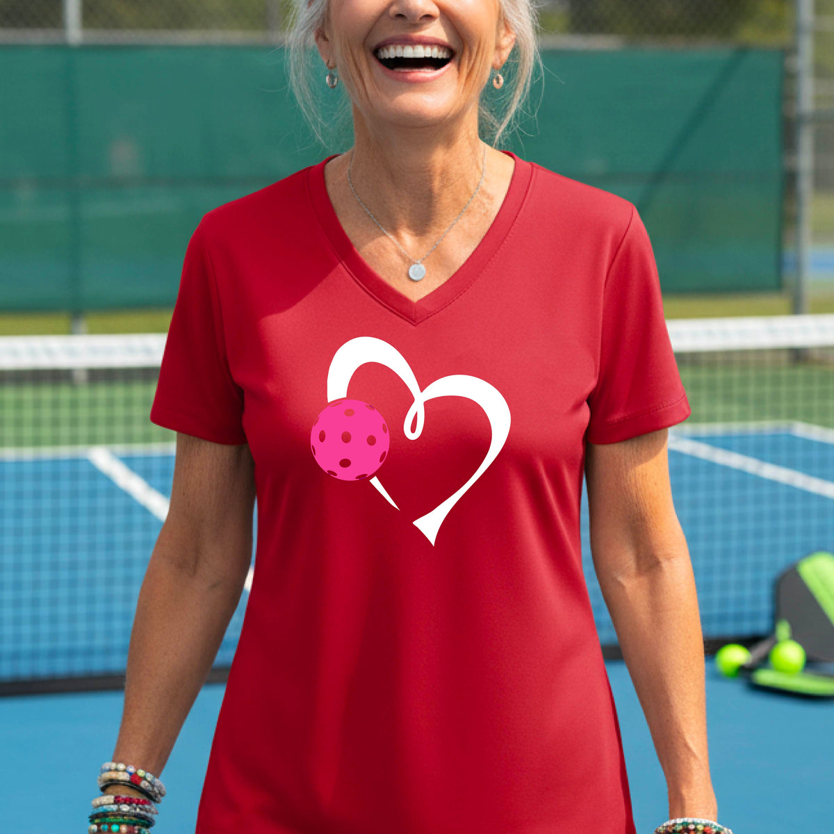 A woman laughing on a pickelball court wearing a red V-neck t-shirt with a white heart outline and a pink pickleball graphic.