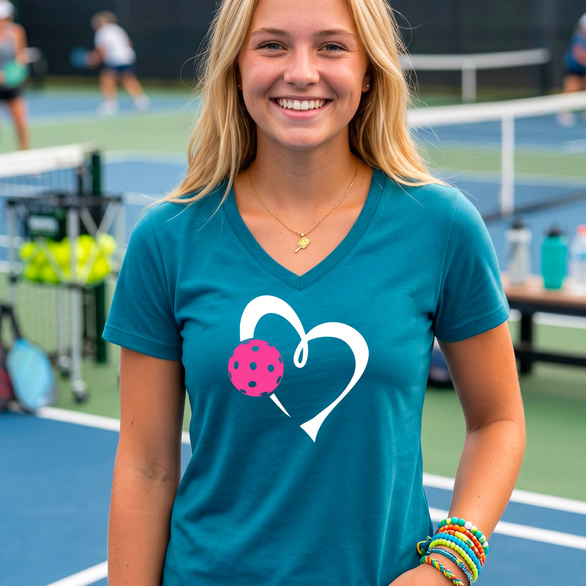 A woman laughing on a pickelball court wearing a teal V-neck t-shirt with a white heart outline and a pink pickleball graphic.