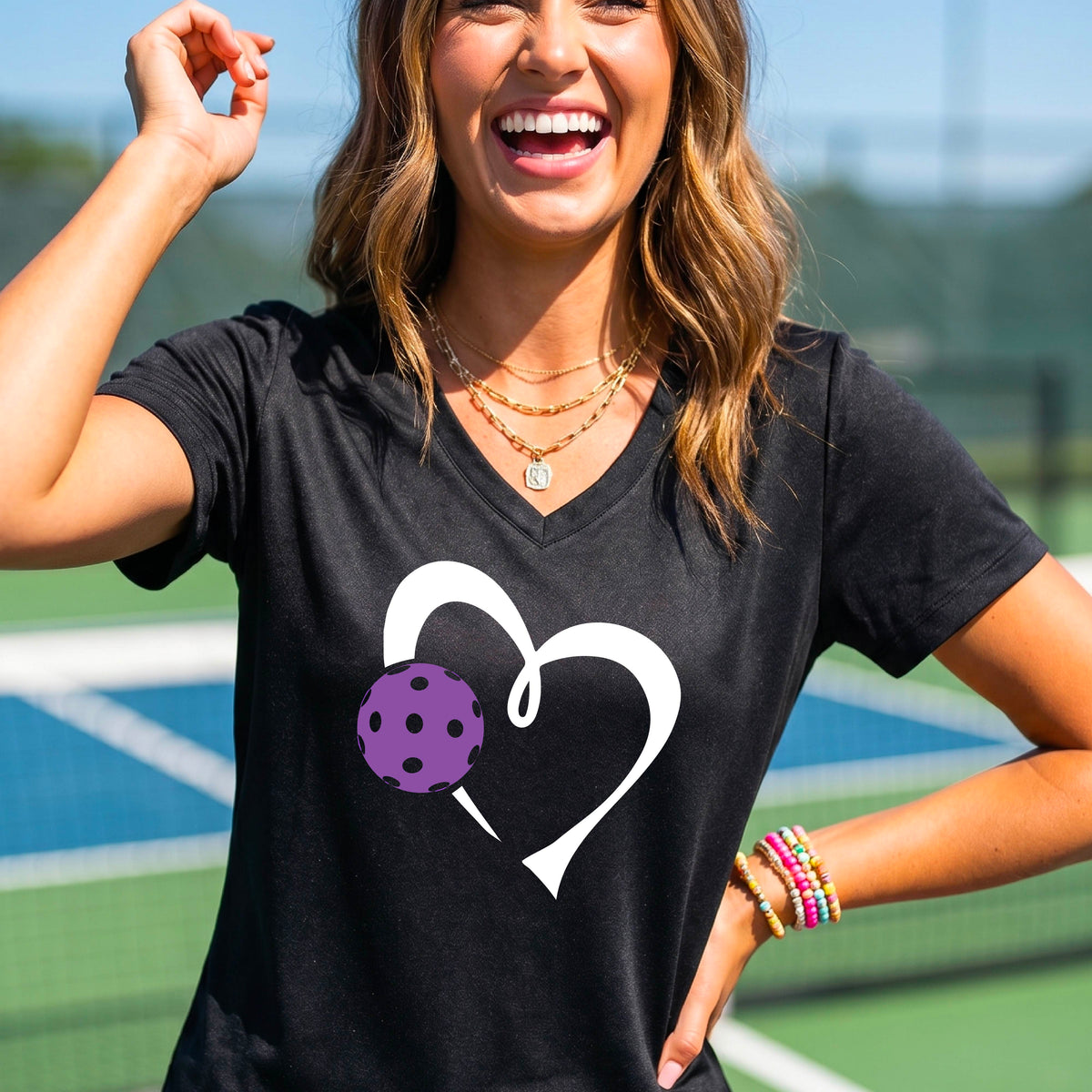 A woman wearing a black V-neck t-shirt with a white heart outline and a purple pickleball graphic, standing on a pickleball court.