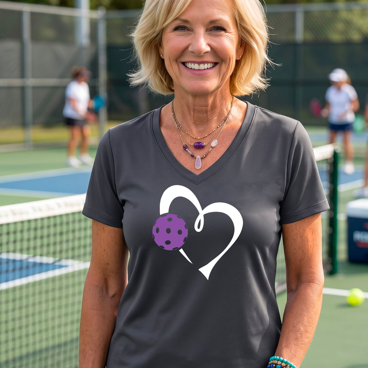 A woman wearing a dark gray V-neck t-shirt with a white heart outline and a purple pickleball graphic, standing on a pickleball court.