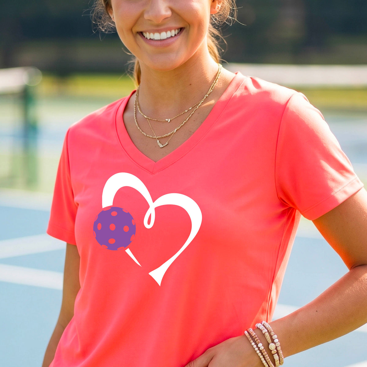 A woman wearing a coral pink V-neck t-shirt with a white heart outline and a purple pickleball graphic, standing on a pickleball court.