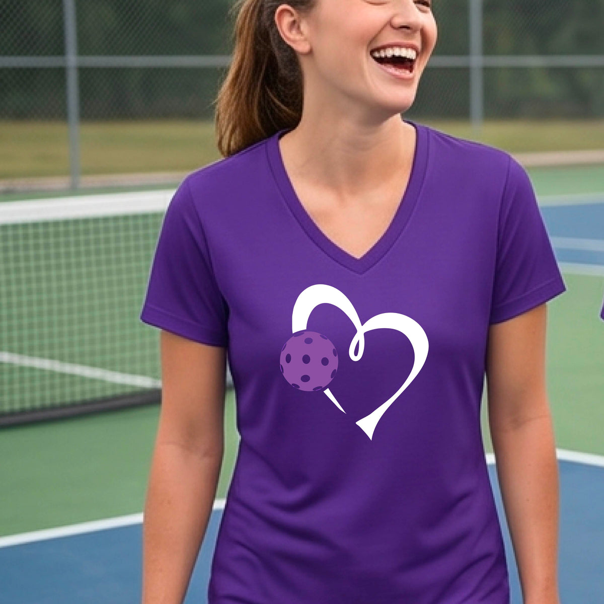 A woman wearing a purple V-neck t-shirt with a white heart outline and a purple pickleball graphic, standing on a pickleball court.