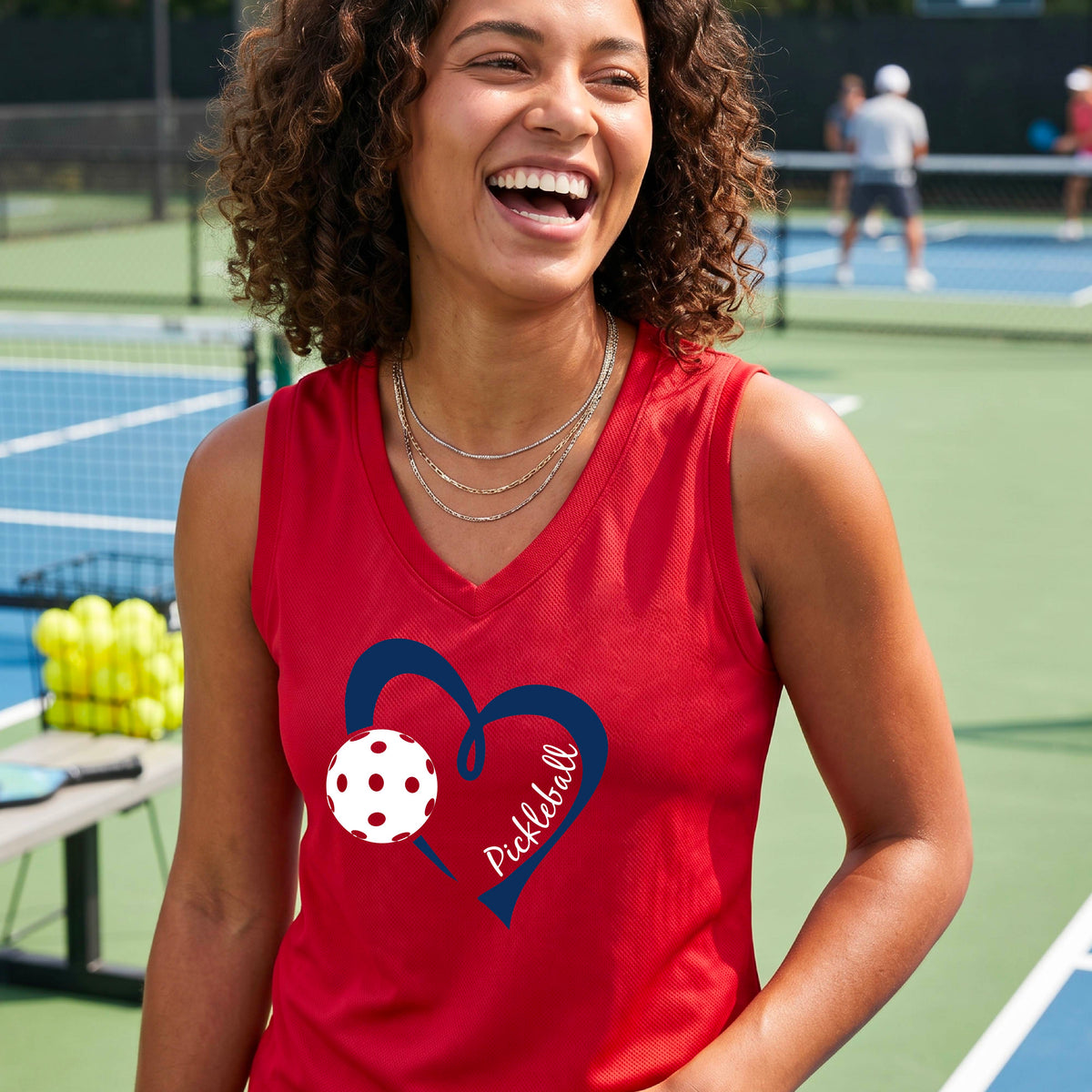 A smiling woman on a pickleball court wearing a red sleeveless tank top with a navy heart outline, a white pickleball graphic, and the word 'Pickleball' printed on the front.