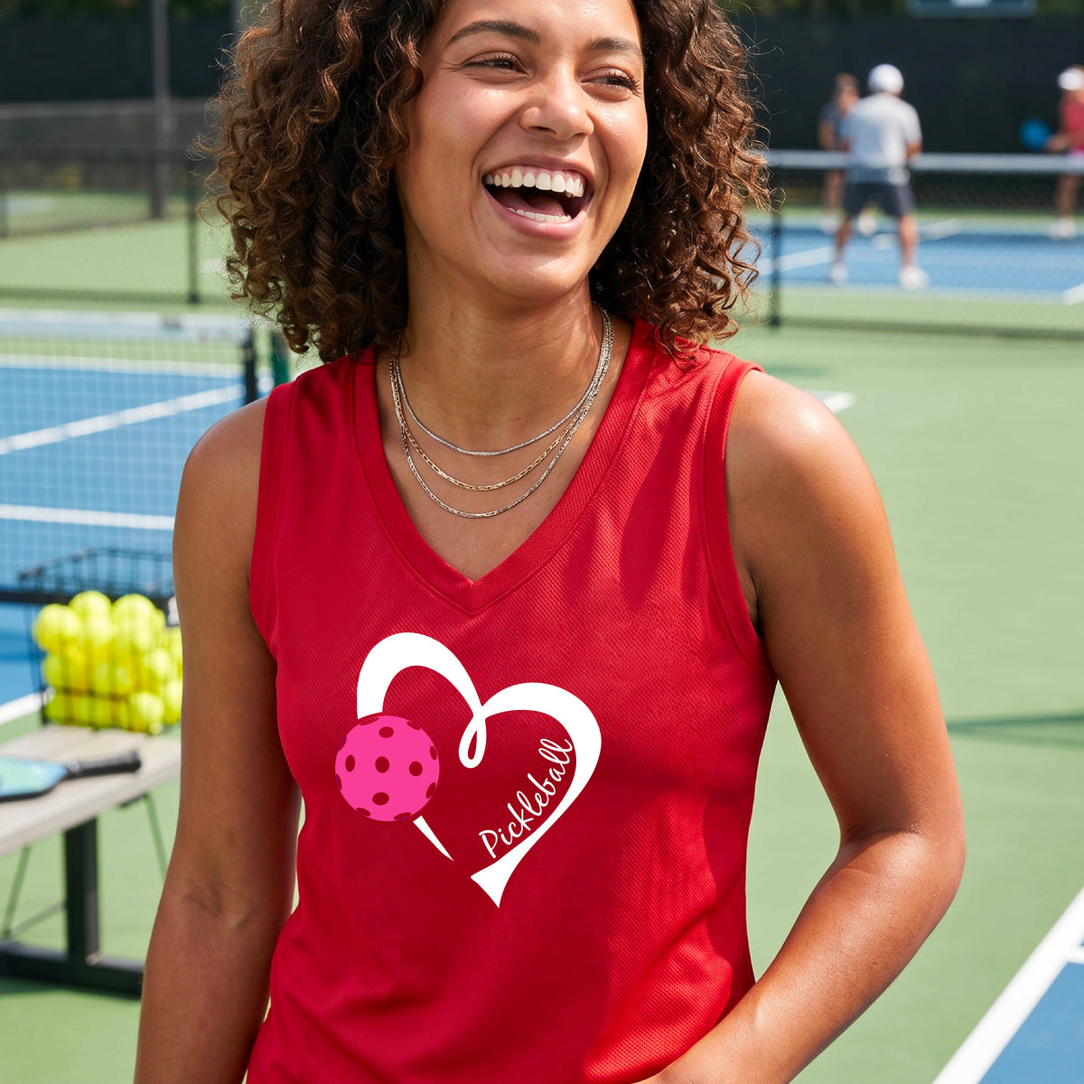 A smiling woman on a pickleball court wearing a red sleeveless athletic tank top with a white heart outline, pink pickleball graphic, and the word 'Pickleball' printed on the front.