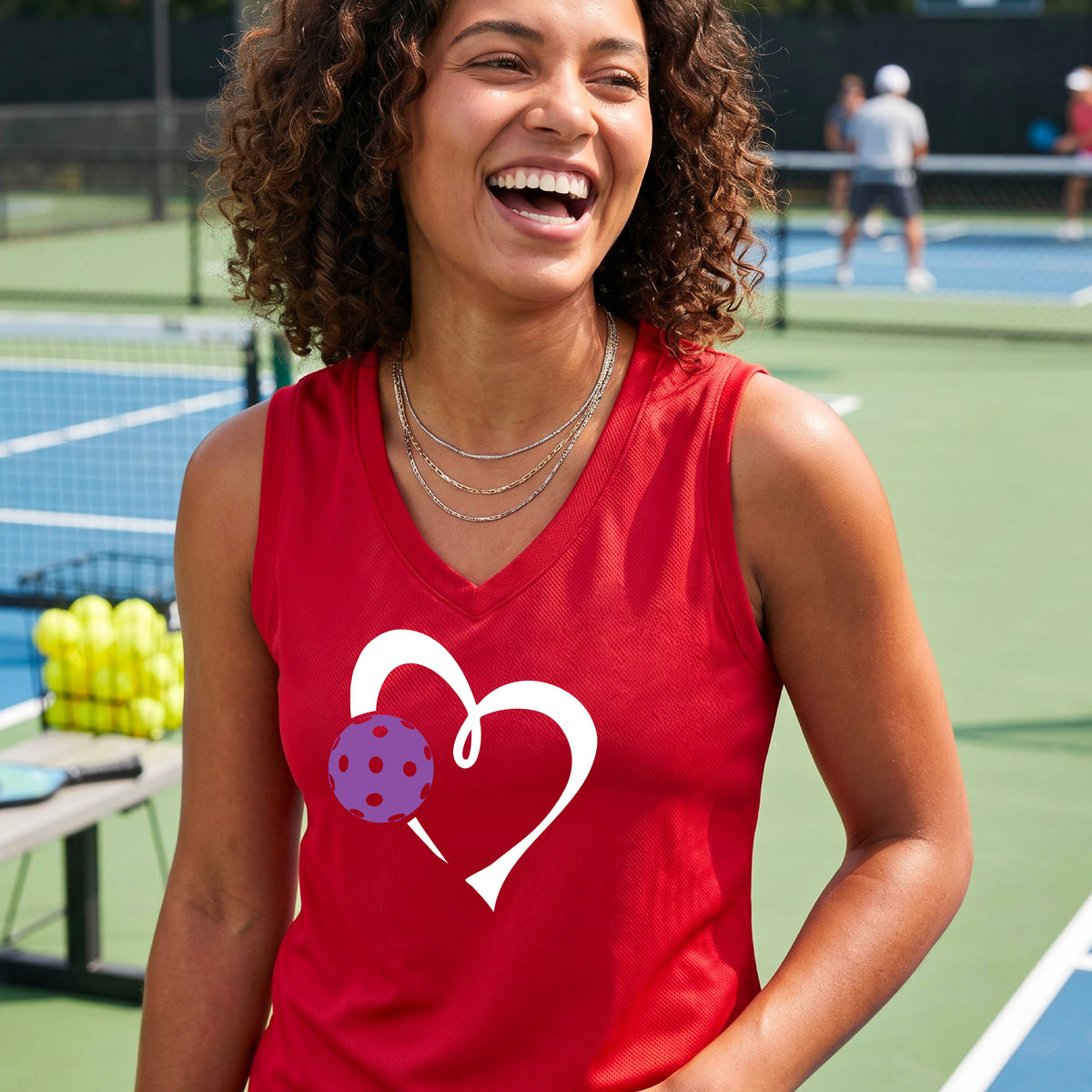 A woman smiling on an outdoor pickleball court wearing a red athletic tank top with a white heart outline and a purple pickleball graphic on the front.