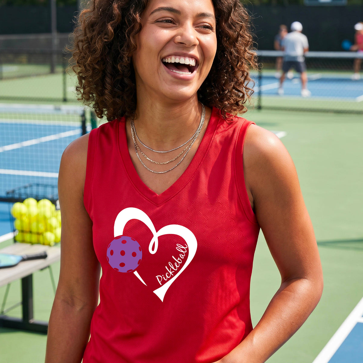 A woman smiling on a pickleball court wearing a red sleeveless tank top with a white heart outline, a purple pickleball graphic, and the word 'Pickleball' printed on the front.