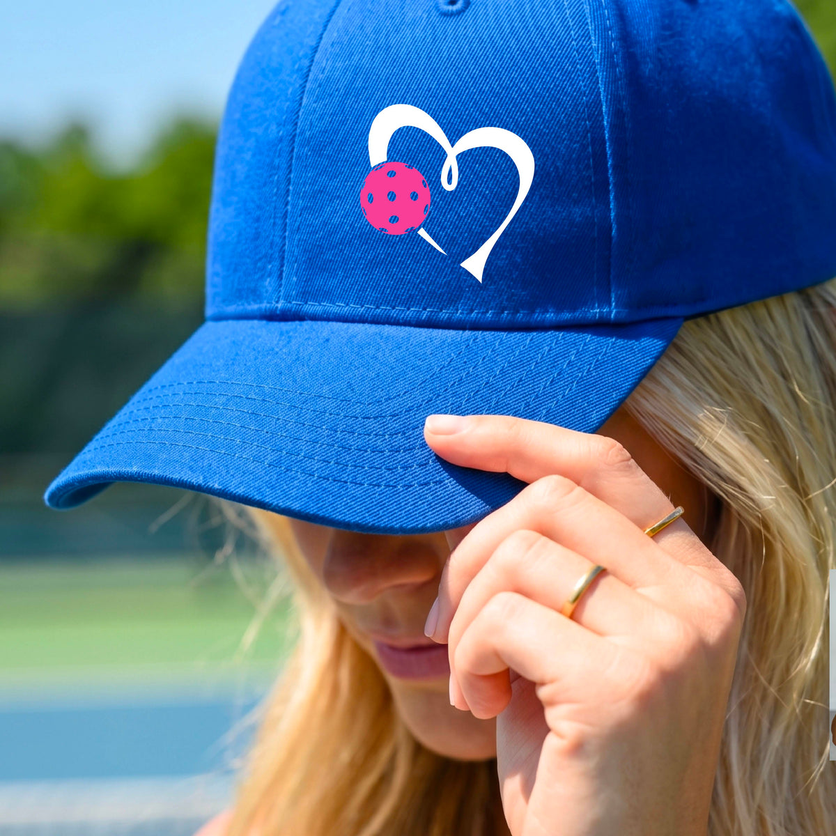 A close-up of a royal blue baseball cap with a white heart outline and a pink pickleball graphic with a blurred outdoor pickleball court background.