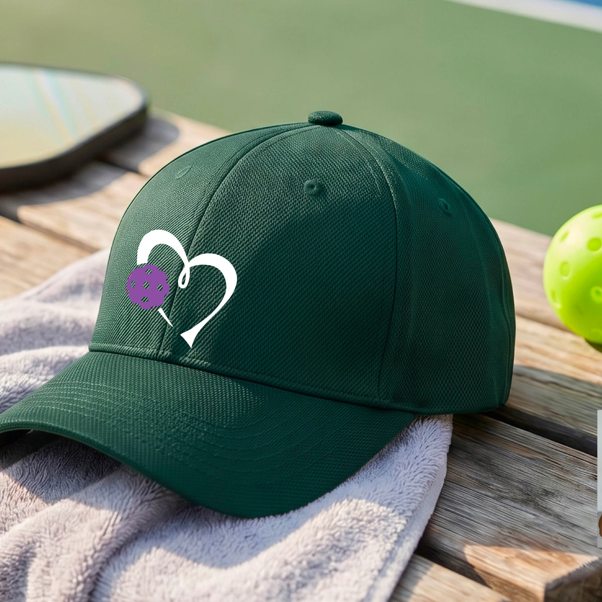 A close-up of a green baseball cap with a white heart outline and a purple pickleball graphic on the front.