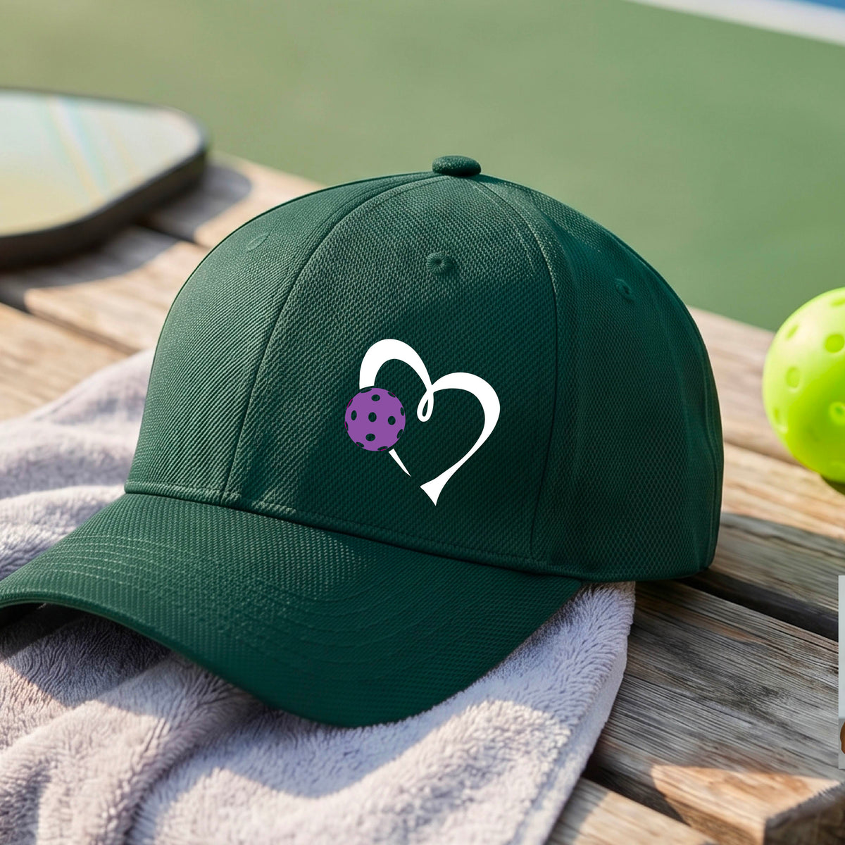 A close-up of a green baseball cap with a white heart outline and a purple pickleball graphic on the front.