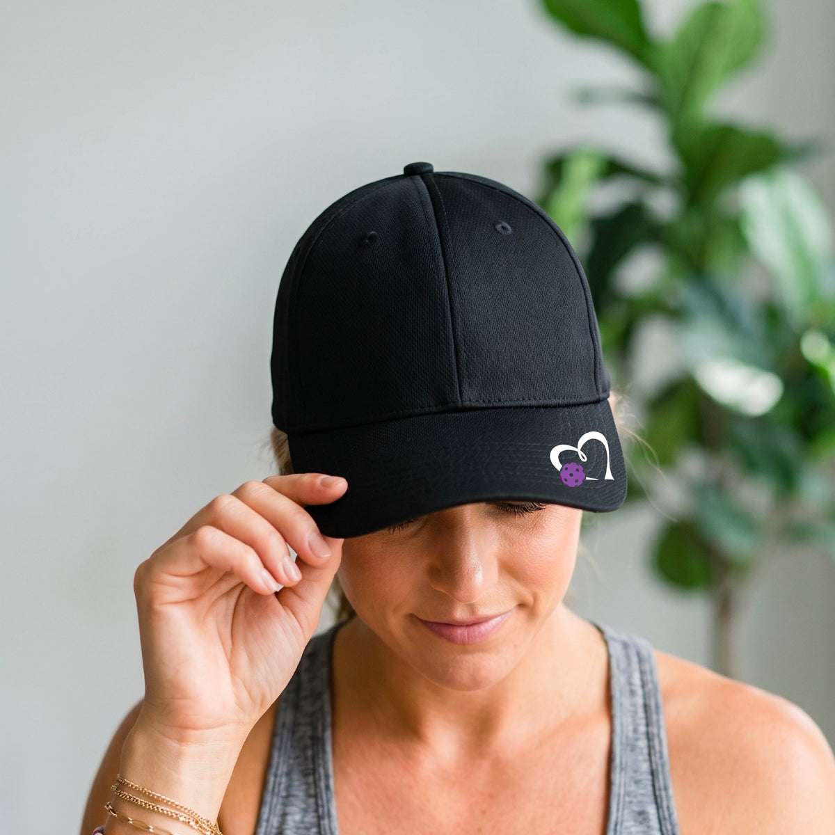 A close-up of a black baseball cap with a white heart outline and a purple pickleball graphic on the front.