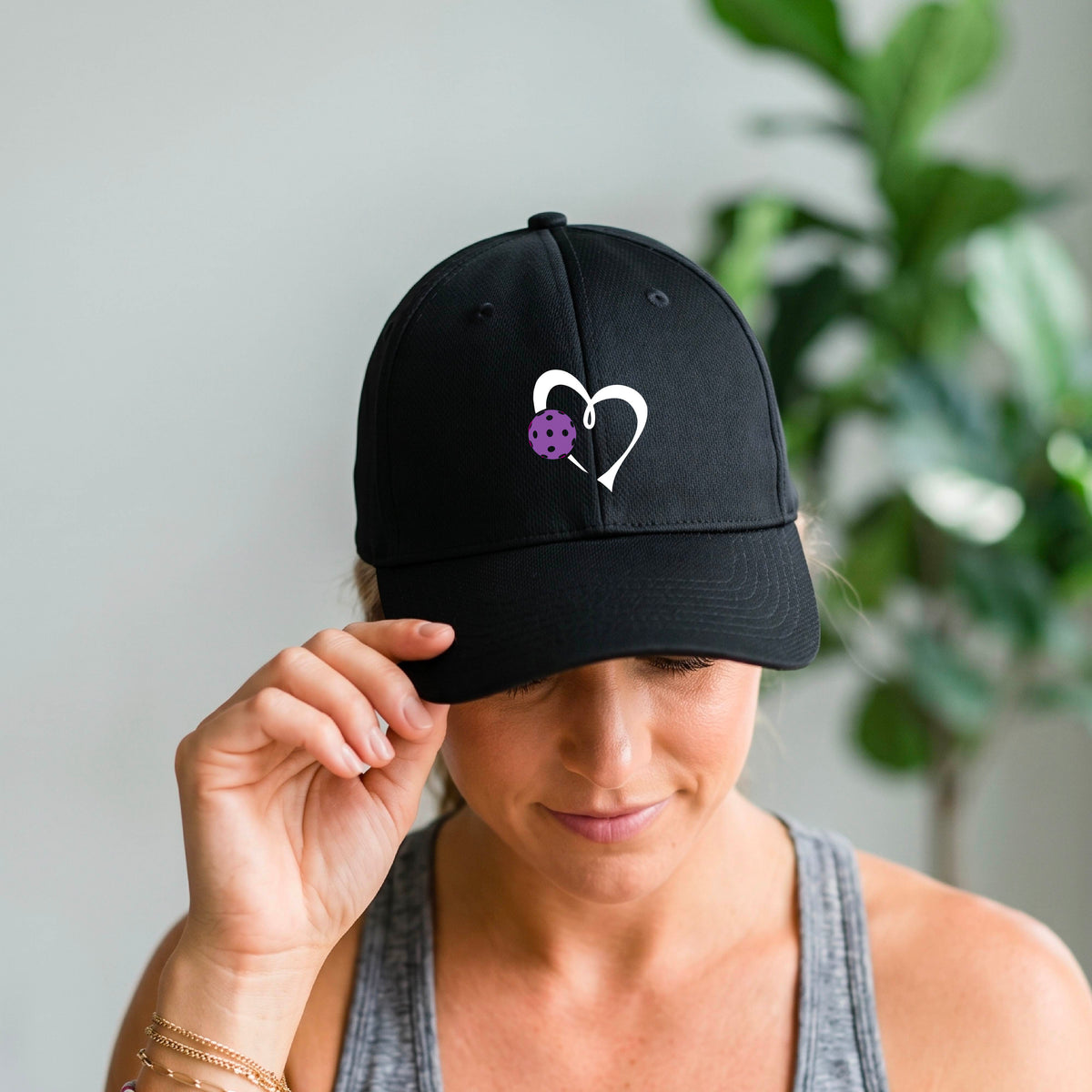 A close-up of a black baseball cap with a white heart outline and a purple pickleball graphic on the front.