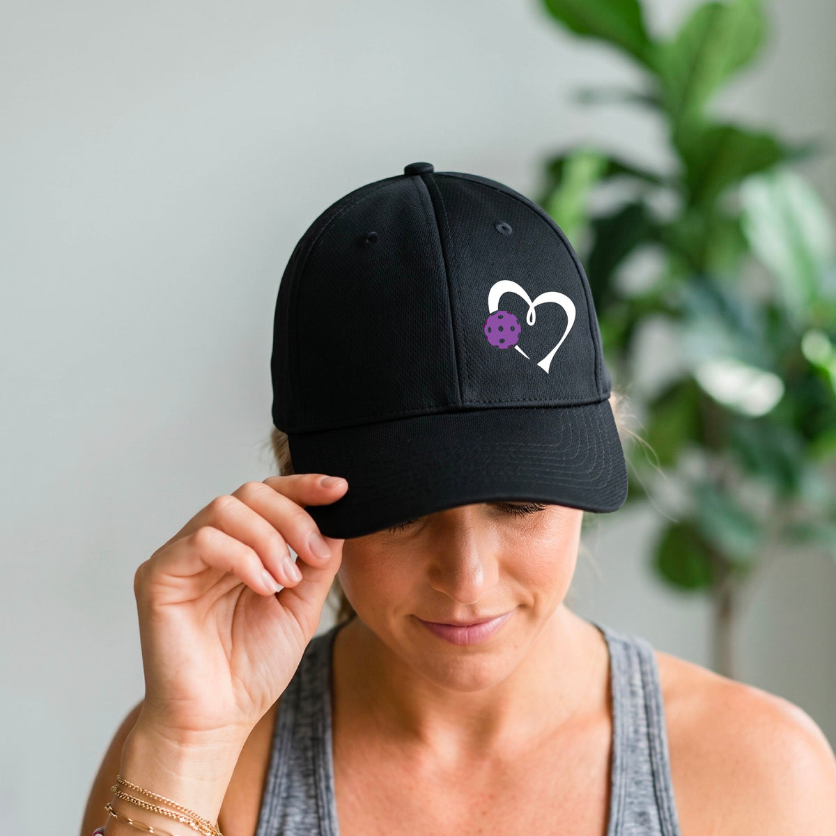 A close-up of a black baseball cap with a white heart outline and a purple pickleball graphic on the front.