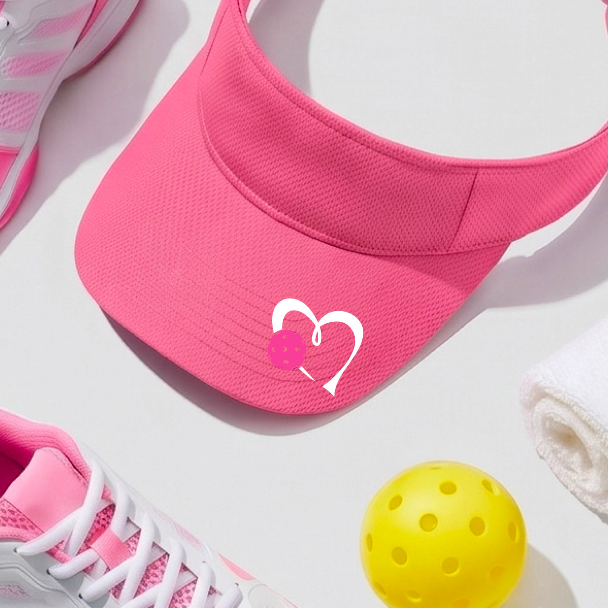 A pink athletic visor with a white heart outline and a pink heart and pickleball graphic on the brim, photographed outdoors on what appears to be a pickleball court.